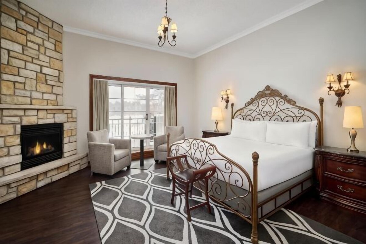 A hotel room with a fireplace, a large bed, a desk with chairs, and a view through a window.