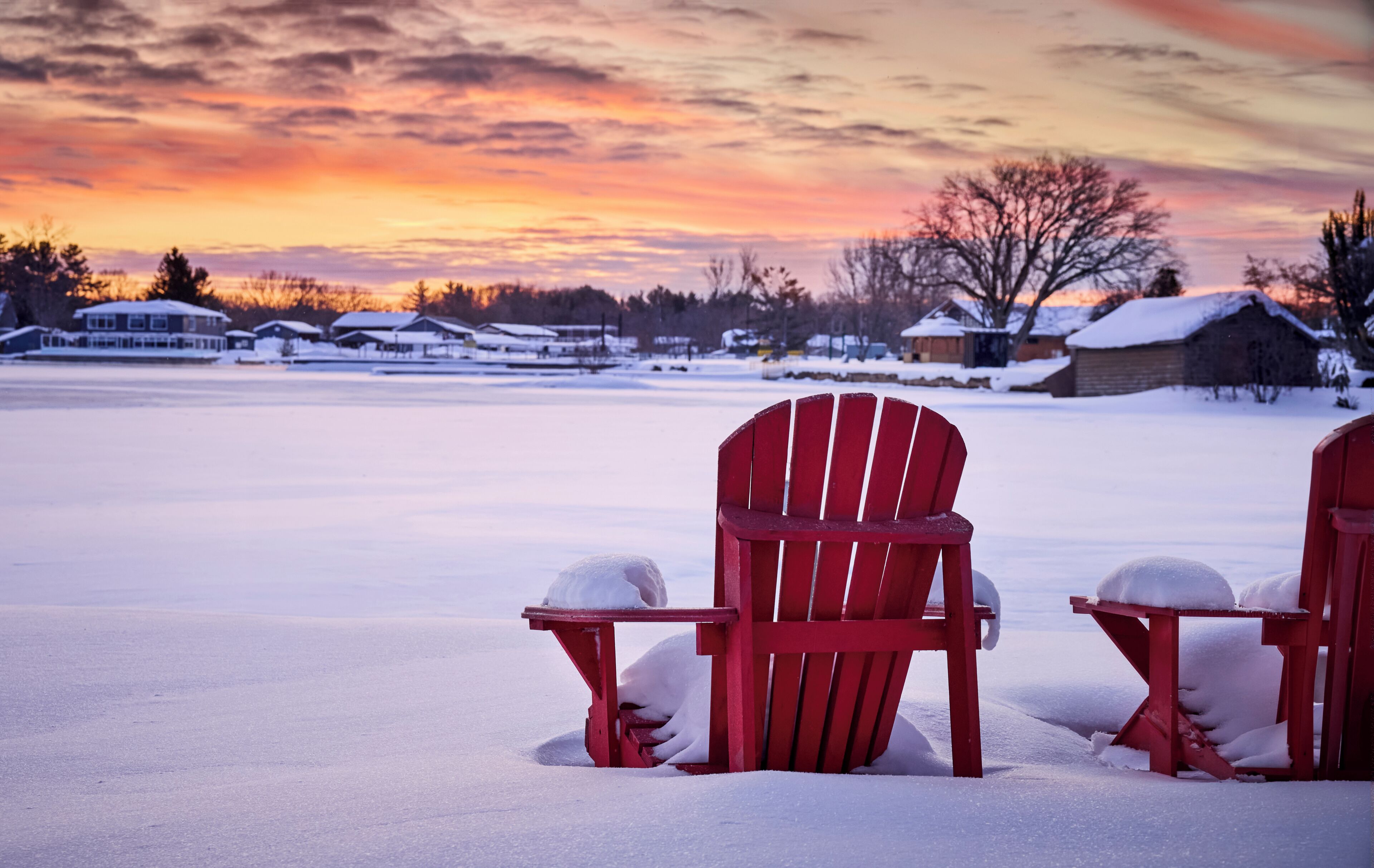 A snow-covered landscape with a sunset, two red Adirondack chairs, and a house in the background.