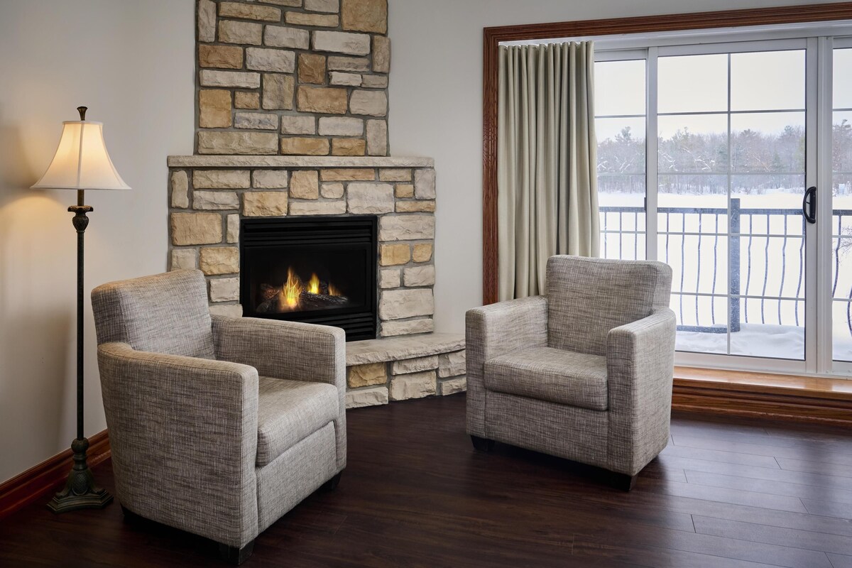 A room with a stone fireplace, two armchairs, a floor lamp, and a view through a large window.