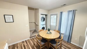 Um quarto com uma mesa redonda de madeira, duas cadeiras, um vaso de planta, uma máquina de lavar, uma porta e uma janela com cortinas.