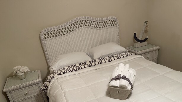 Una cama bien hecha con colcha blanca, almohada estampada y una pequeña cesta de toallas dobladas sobre una mesita de noche de mimbre blanco.