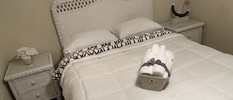 Una cama bien hecha con colcha blanca, almohada estampada y una pequeña cesta de toallas dobladas sobre una mesita de noche de mimbre blanco.