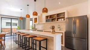 A modern kitchen with a central island, bar stools, a stainless steel refrigerator, pendant lights, and a view to the outside. - 5-Bed Deluxe Condo • Private Villa Feel • Full Hotel Amenities (Tulum)