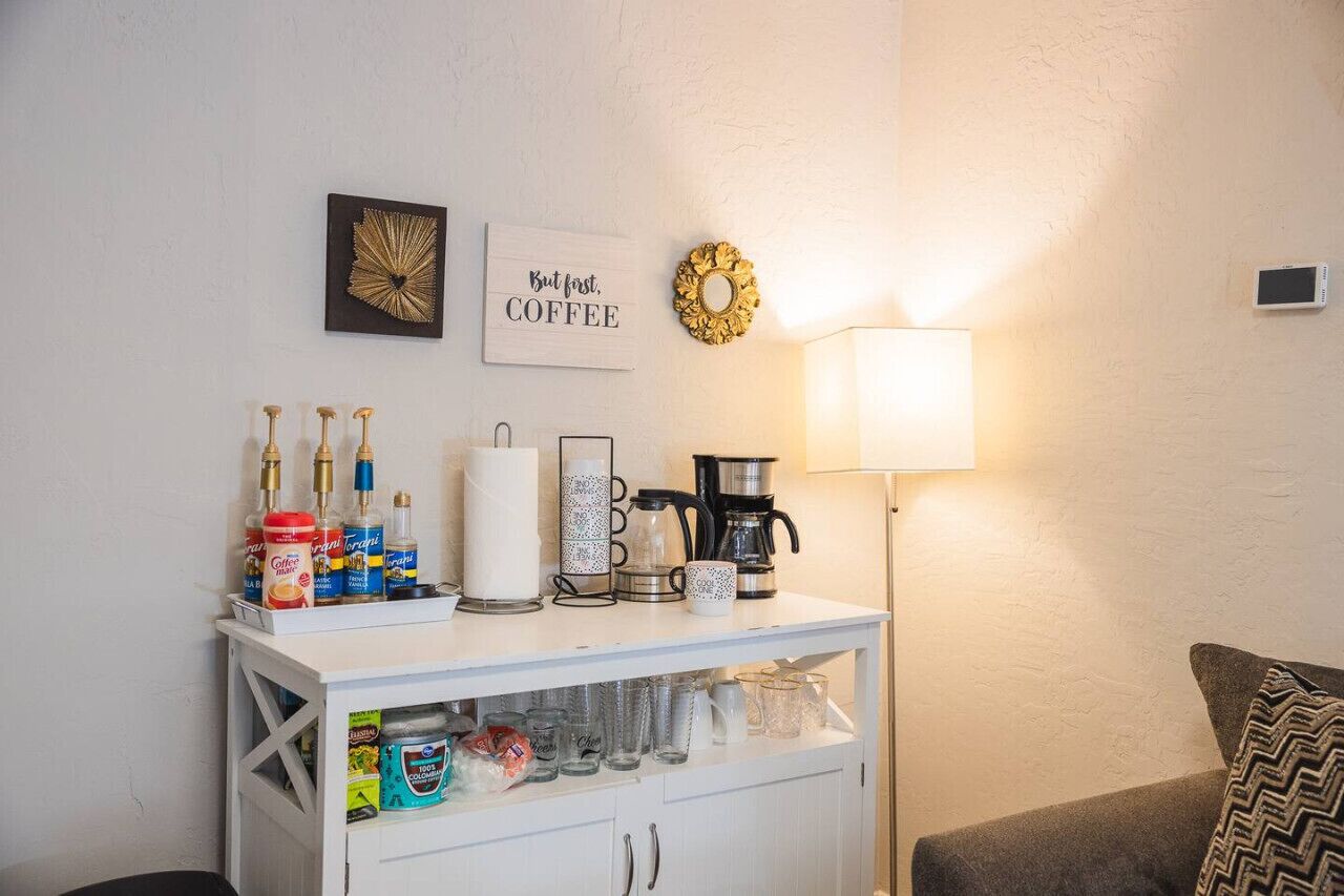 Una kitchenette con equipo para preparar café, un mueble blanco con espacio de almacenamiento y una decoración de pared con temática cafetera.
