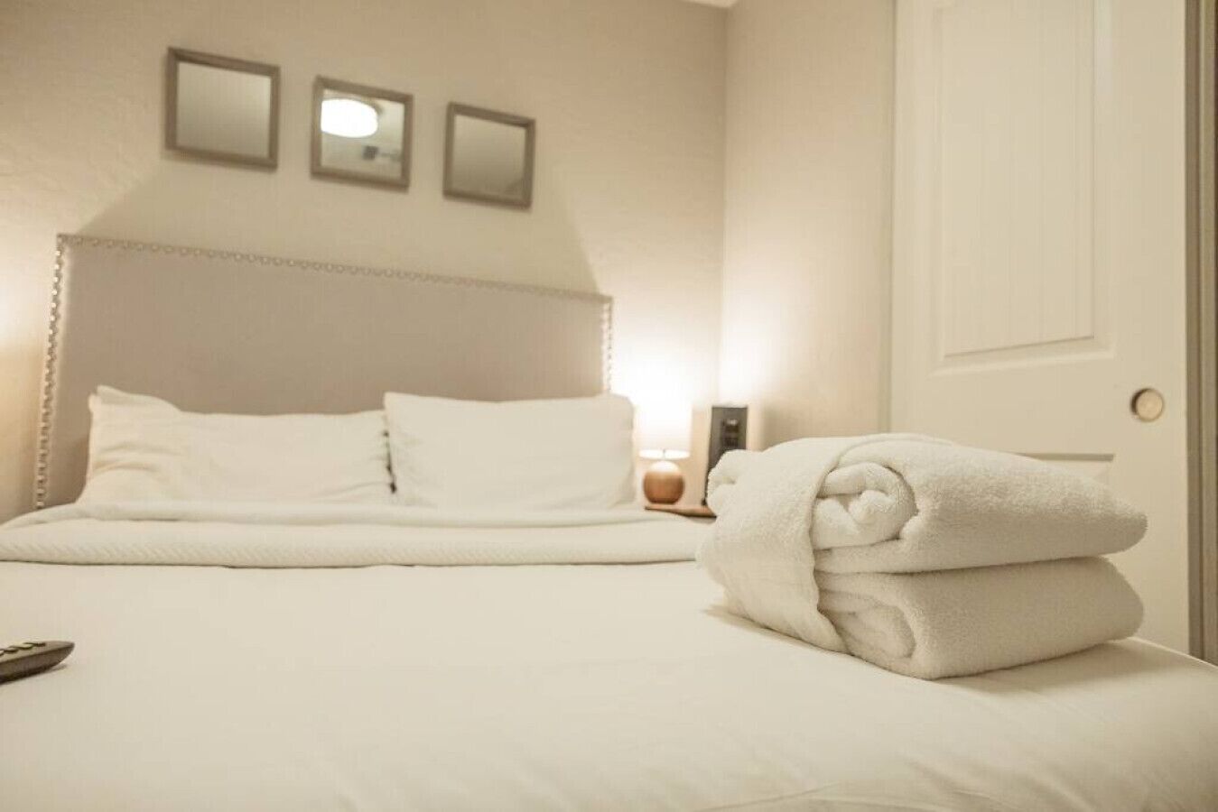 Un lit bien fait, avec du linge de lit blanc et des oreillers, une serviette pliée posée à côté, et trois tableaux encadrés accrochés au mur.