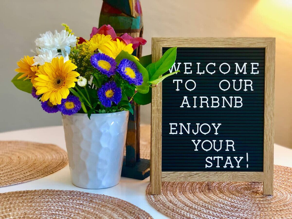 A welcome sign for an Airbnb with a message 'WELCOME TO OUR AIRBNB ENJOY YOUR STAY!' next to a vase of colorful flowers.