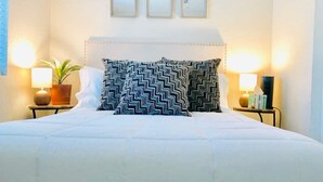 Un letto rifatto con un copriletto bianco, due cuscini a motivi bianchi e neri e una testiera con decorazioni. Sopra il letto, sulla parete, ci sono tre quadri incorniciati.