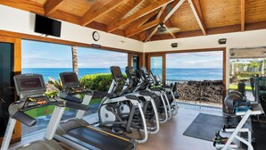 A gym with treadmills and elliptical machines, large windows with a view of the ocean, and a wooden ceiling.