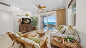 A modern living room with a television, wooden furniture, a ceiling fan, and a view of the ocean through large windows.