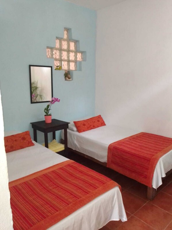 A room with two beds, each with a red-striped bedspread, a small table with a potted plant, and a wall-mounted light fixture.