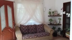 En stue med en beige sofa, en loftventilator, lette gardiner og potteplanter.