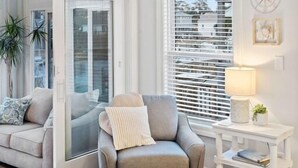 Una sala de estar con un sillón gris, un sofá, un tapete redondo, una mesita auxiliar blanca y una ventana con persianas.