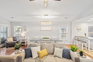 A spacious living room with a grey sofa, a dining table, and a kitchen area with white bar stools.