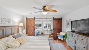 Un dormitorio con una cama, ventilador de techo, televisor, cómoda y un ventanal grande.