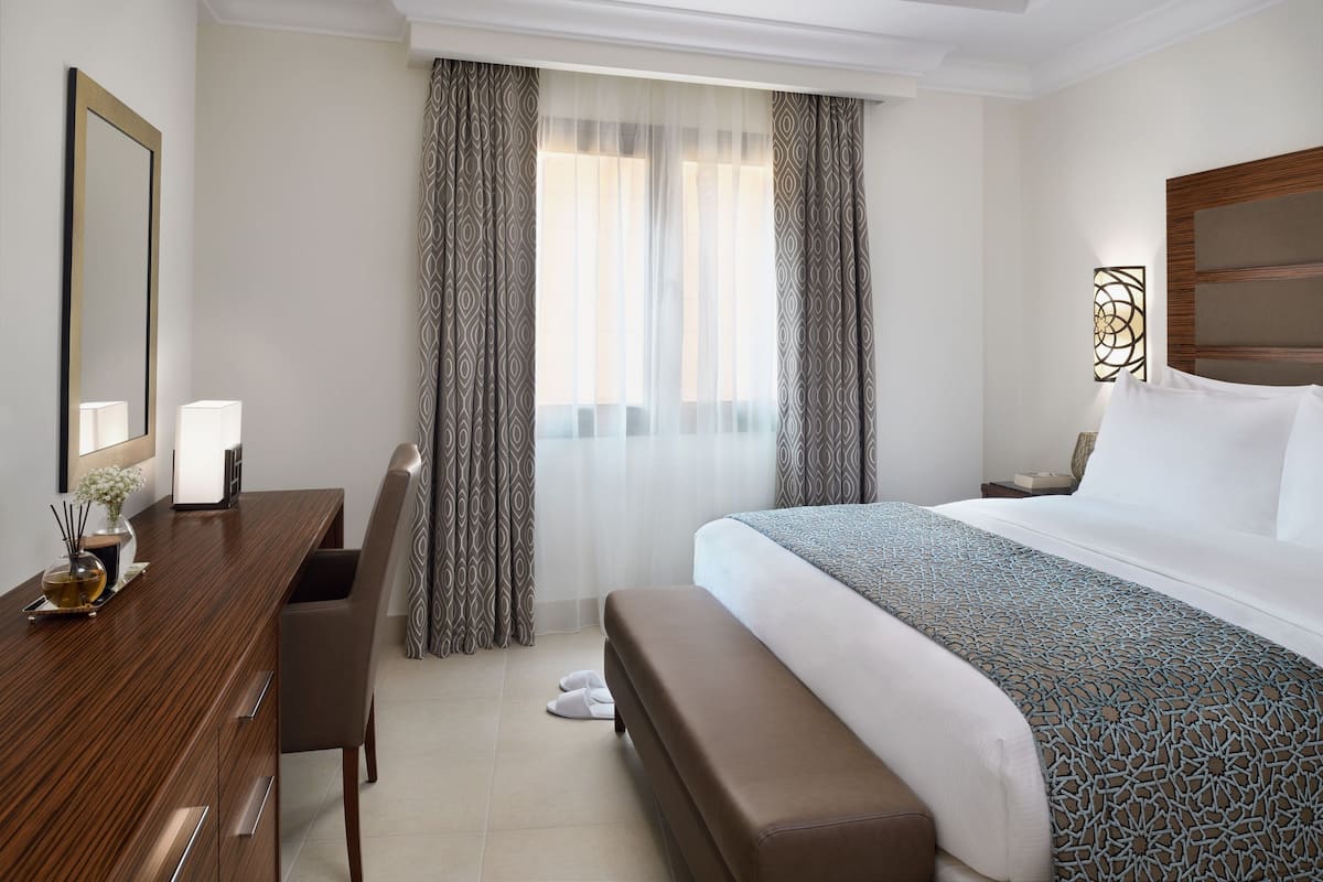A hotel room with a wooden desk, a bed with a patterned bedspread, a bench, and sheer curtains.