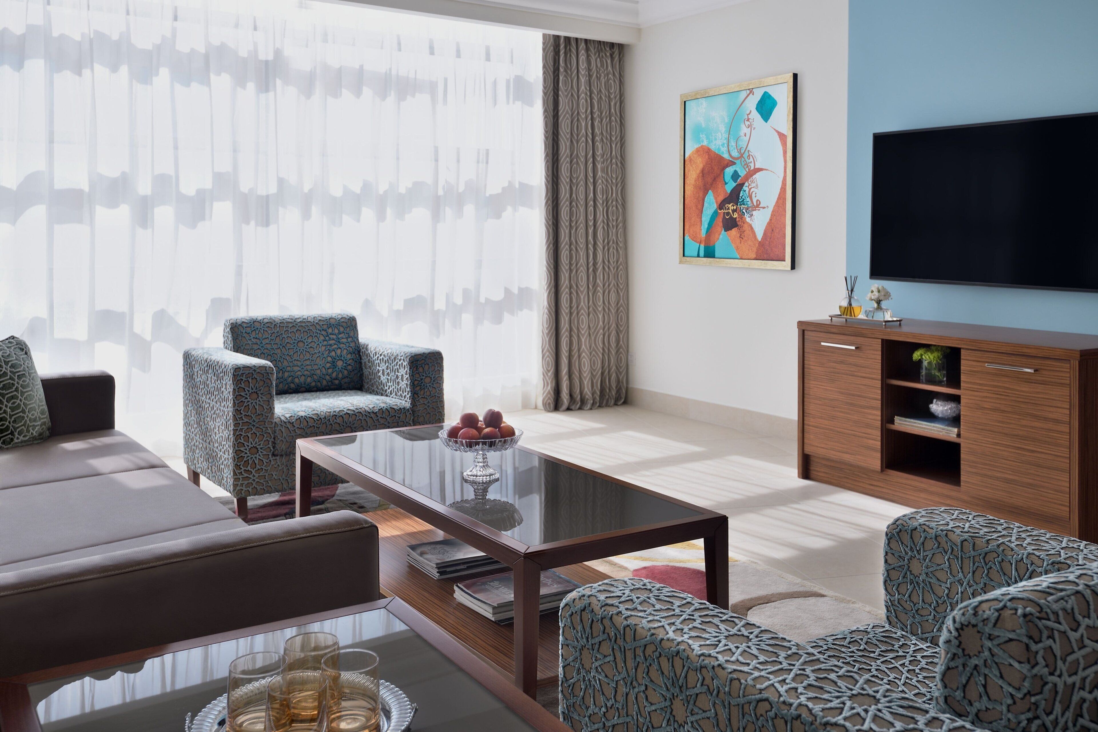 A modern living room with a sofa, patterned armchairs, a glass coffee table, a wooden cabinet, and a flat-screen TV.