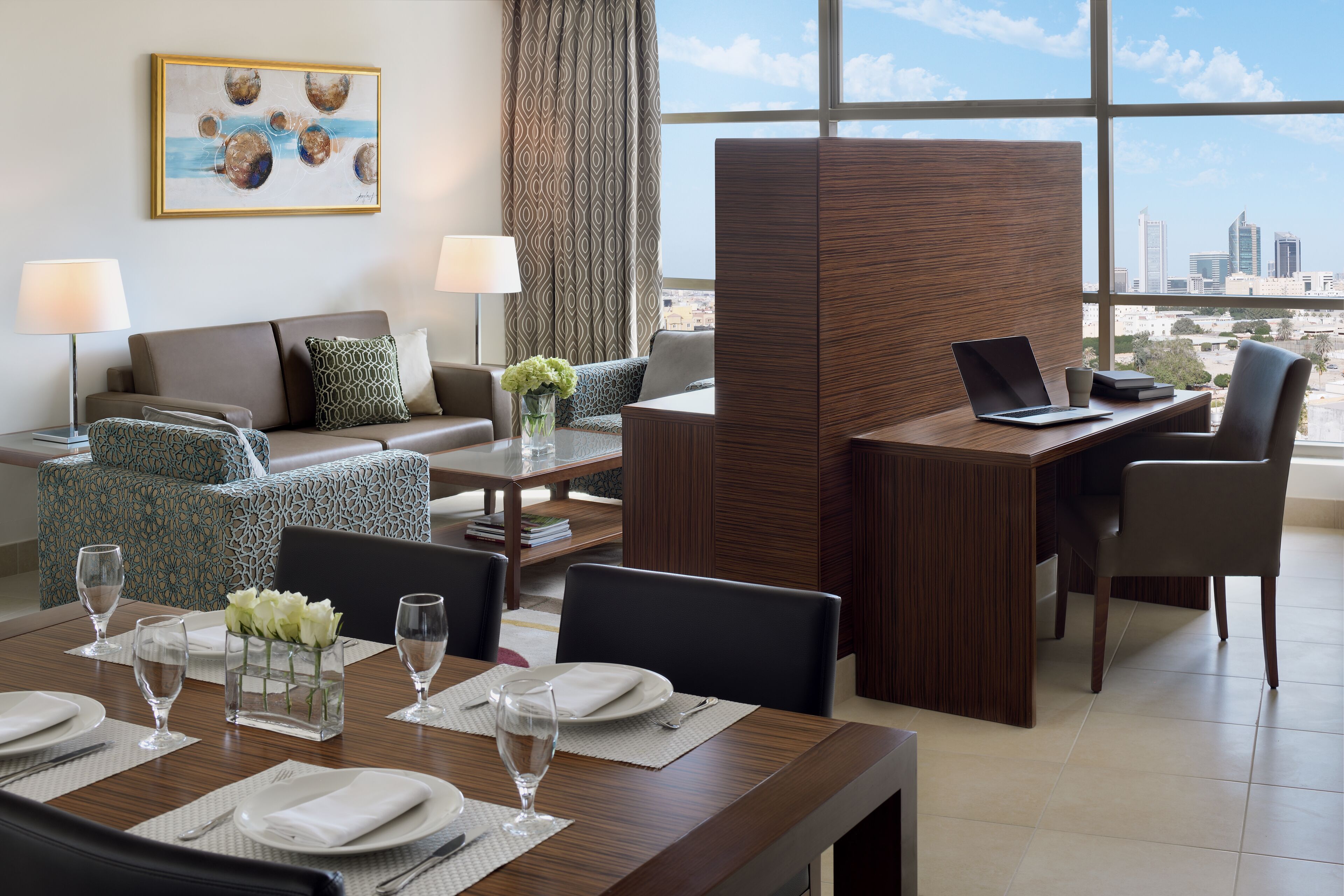 A modern hotel room with a dining area, a sofa, a desk, and a view of the city skyline.