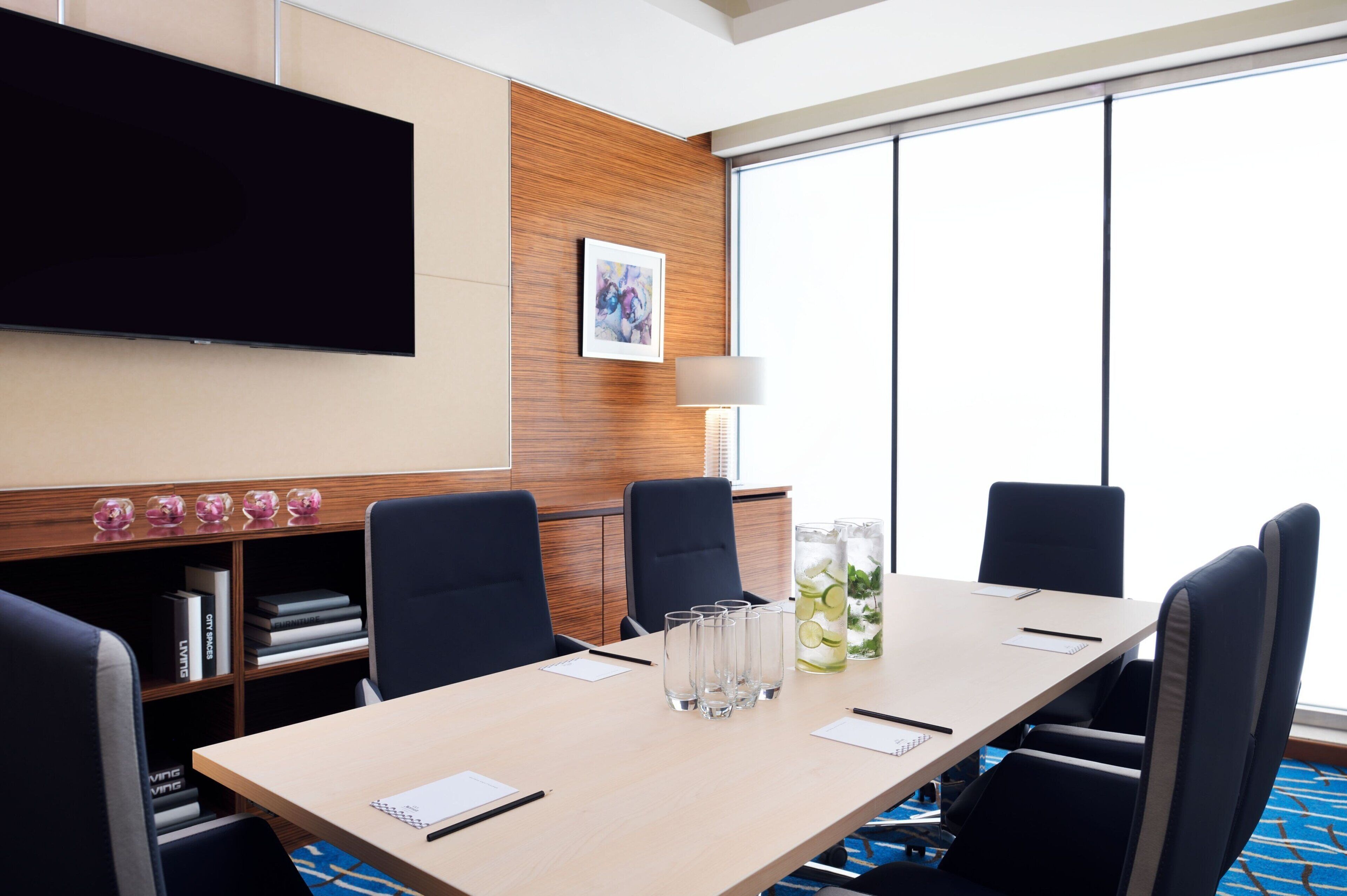 A conference room with a long table, four chairs, a flat-screen TV, a framed picture, a lamp, and a bookshelf with books.