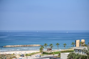 A coastal view with a beach, palm trees, and a modern building with a unique architectural design.