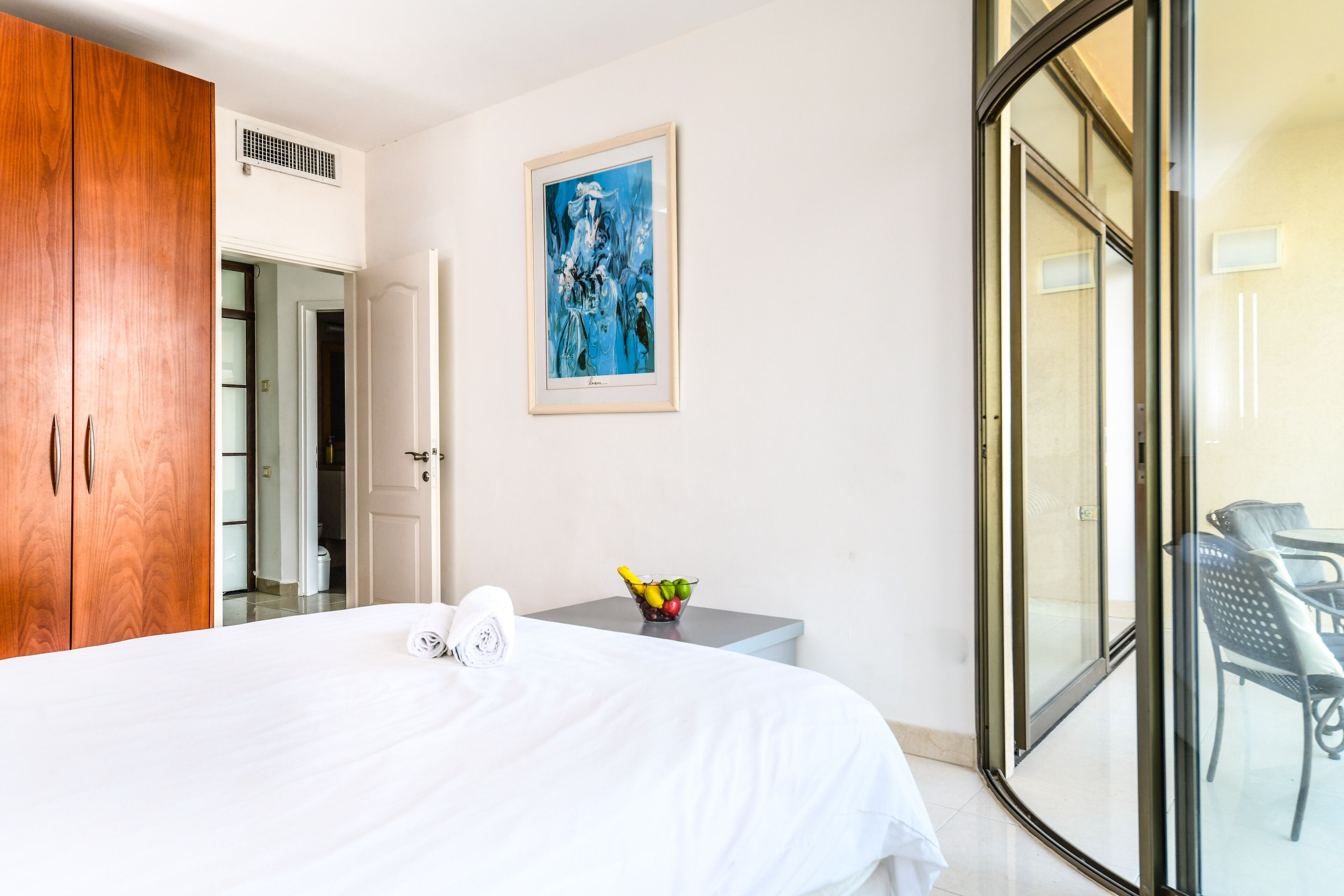A hotel room with a bed, a wooden wardrobe, a framed artwork on the wall, a small table with a fruit bowl, and a sliding glass door leading to a balcony with outdoor furniture.