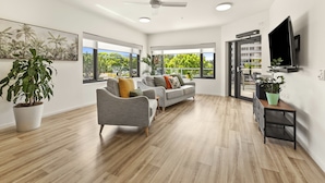 A spacious living room with a grey sofa, a coffee table, and a TV mounted on the wall. There are large windows providing a view of the outdoors. - Healthy Living Centre (Cairns North)