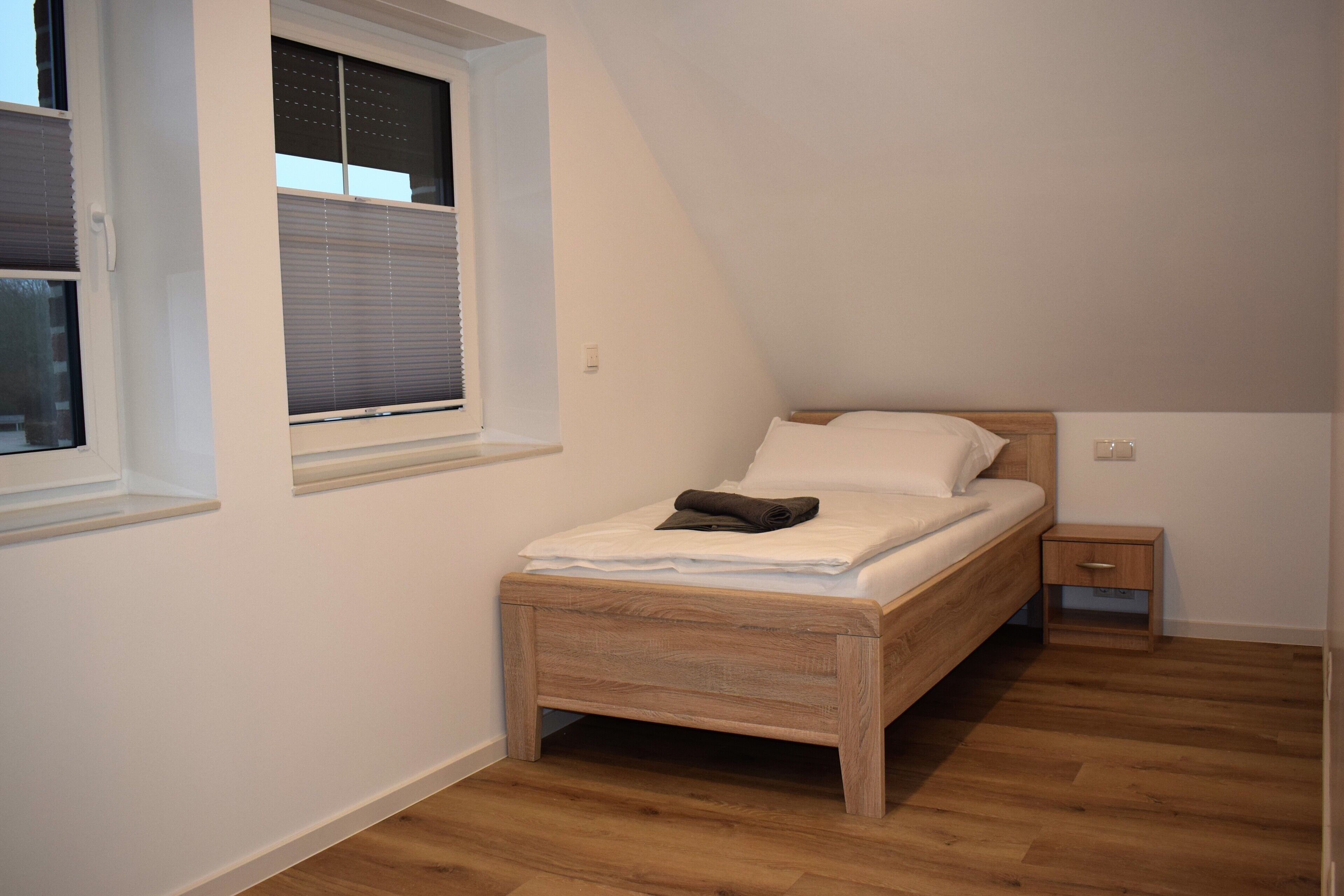 Une chambre avec un lit en bois, une petite table de chevet, une fenêtre avec des stores et un plancher en bois.