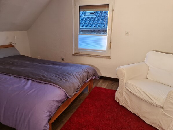 A bedroom with a bed, a window with blinds, a red rug, and a white chair.