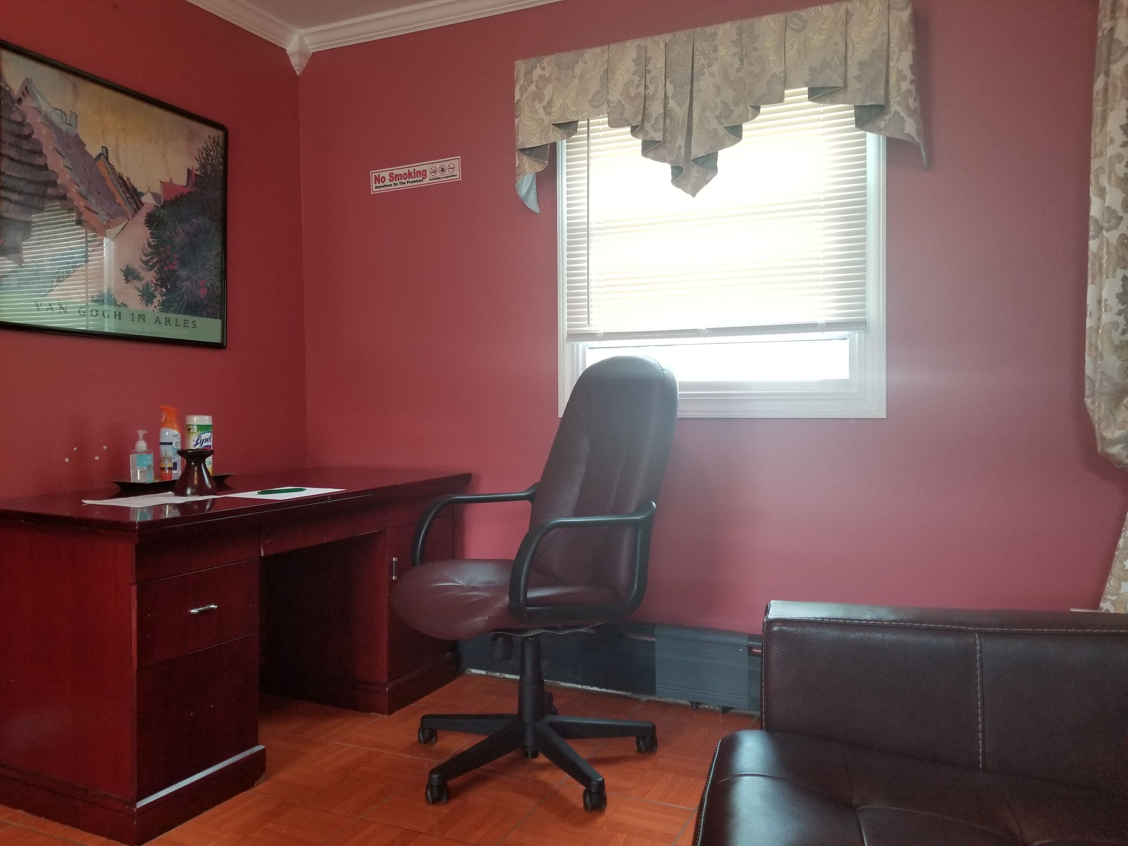 A room with a desk, office chair, and a sofa. There is a window with blinds and a painting on the wall.
