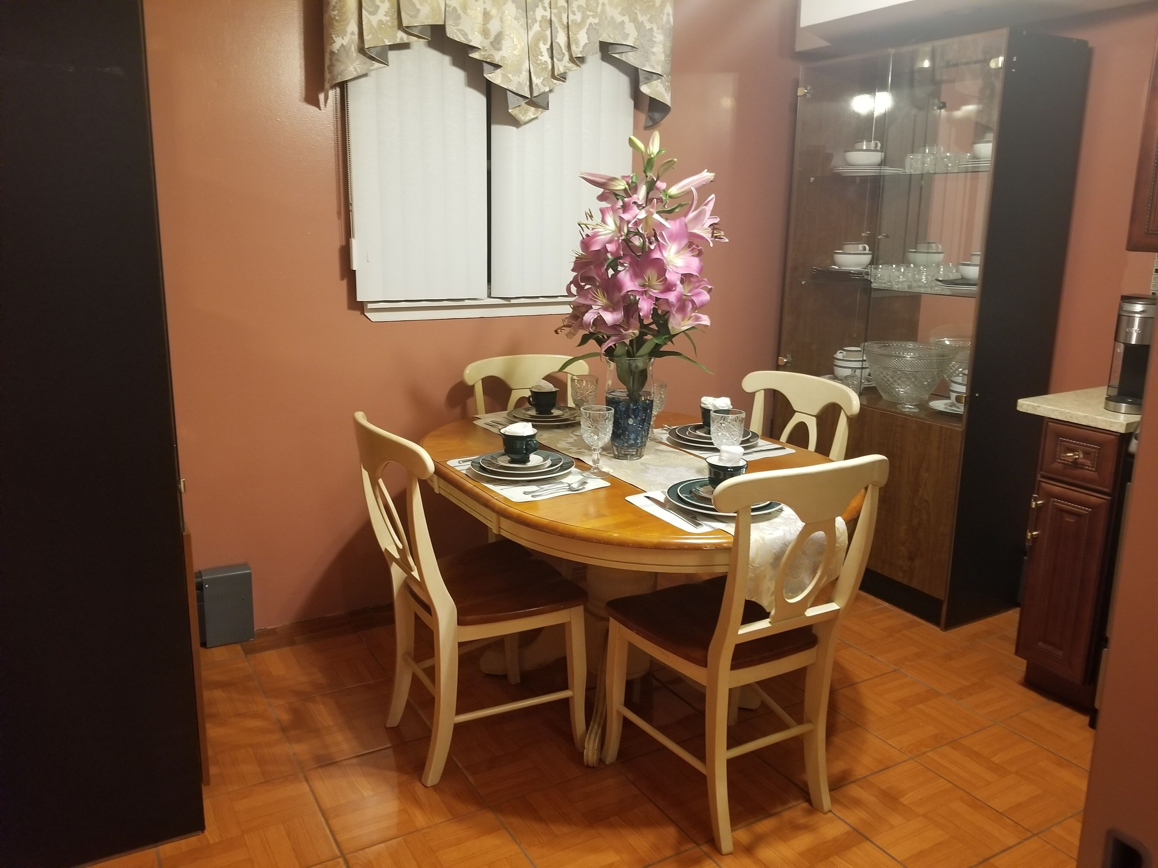 A dining area with a wooden table set for four, surrounded by white chairs. A vase with pink flowers is on the table.