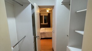 Ein schmaler Flur, der in ein Schlafzimmer mit Bett, Deckenventilator und einem mit Vorhängen versehenen Fenster führt.