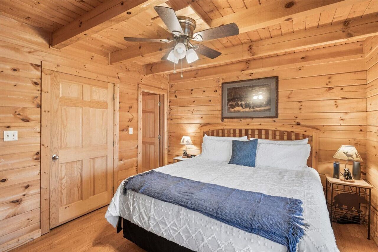 Une chambre à coucher dans une cabane en bois, avec un lit, un ventilateur de plafond, une table de chevet avec une lampe et un tableau encadré au mur.