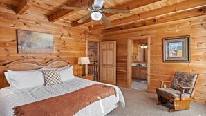 Un dormitorio tipo cabaña de madera con una cama, una silla, un ventilador de techo y una puerta que da a un baño.