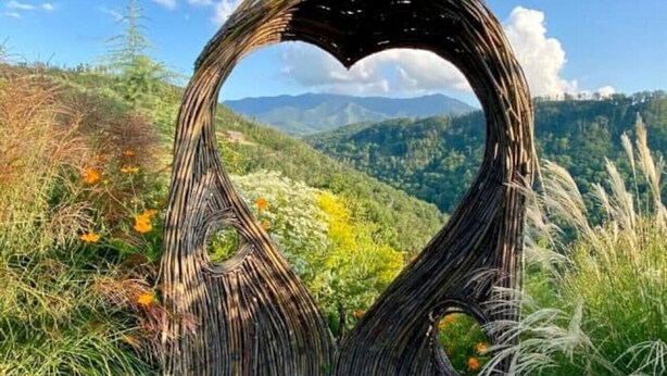 Una estructura de madera con forma de corazón, con un banco de piedra en primer plano, rodeada de vegetación y montañas al fondo.