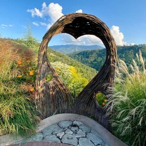 A heart-shaped wooden structure with a stone bench in front, surrounded by greenery and mountains in the background.