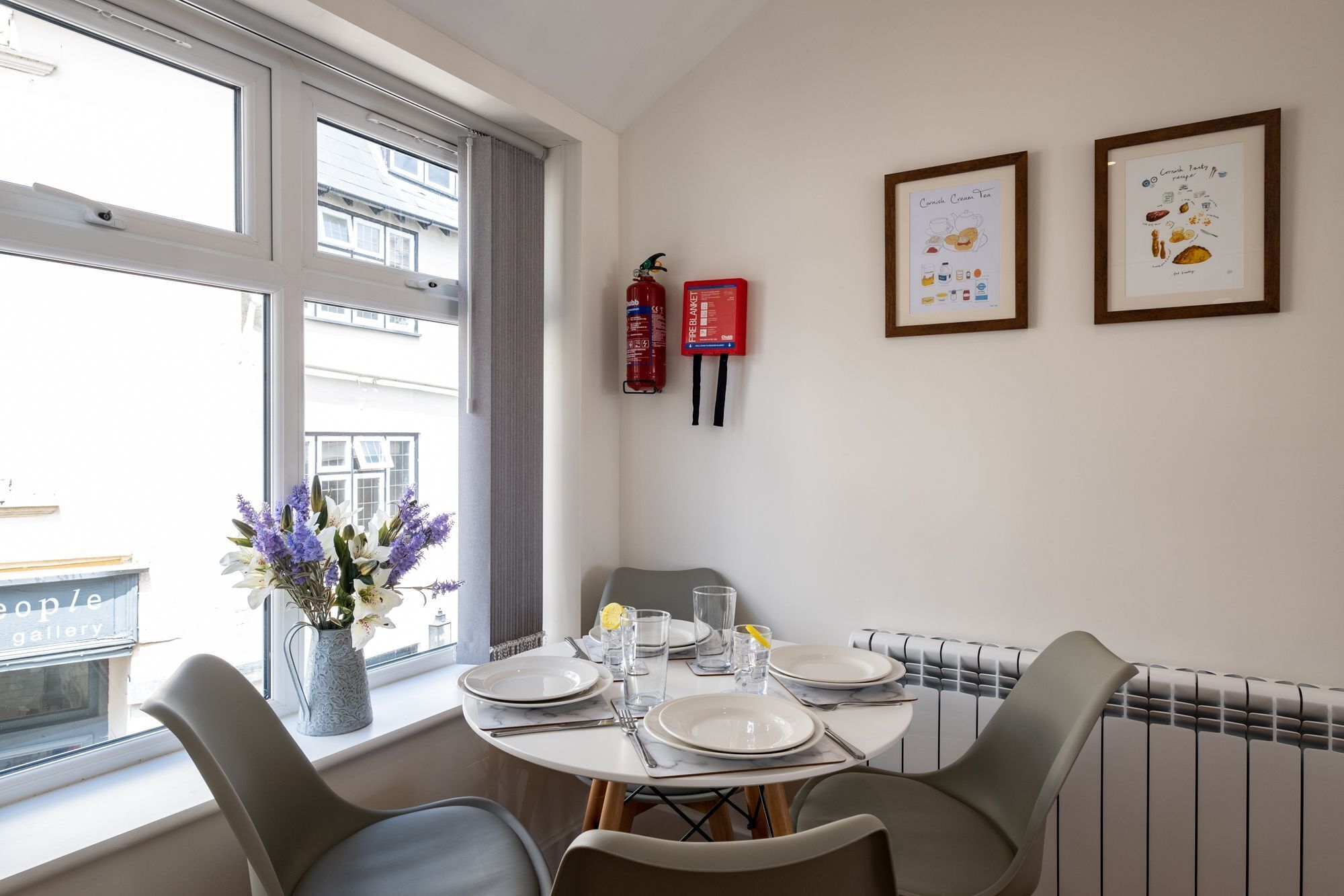 A dining area with a table set for four, a vase of flowers, a fire extinguisher, and two framed pictures on the wall.