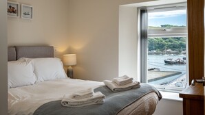 Ein Hotelzimmer mit Bett, Nachttisch, Lampe, Fenster mit Blick auf einen Hafen und zwei gerahmten Bildern an der Wand.