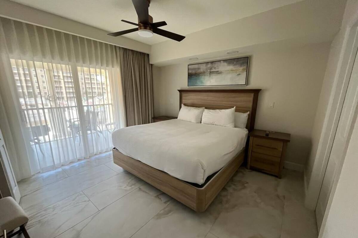 A large bed with a wooden headboard, a nightstand, and a ceiling fan in a hotel room with a view of buildings through sheer curtains.