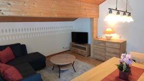Een gezellige woonkamer met houten plafond, een flatscreen televisie, een houten kast, een eettafel met een bloemenvaas, en een blauwe bank met rode kussens.