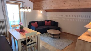 Een gezellige woonkamer met een houten plafond, een donkerblauwe bank met rode kussens, een ronde salontafel, en een eethoek met een houten tafel en stoelen.