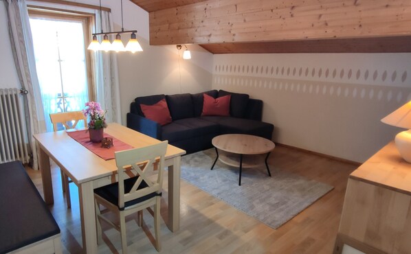 A cozy living room with a wooden ceiling, a dark blue sofa with red cushions, a round coffee table, and a dining area with a wooden table and chairs.