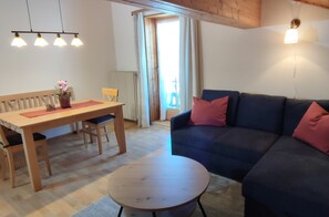A cozy living room with a wooden dining table, a round coffee table, a blue sofa with red cushions, and a wooden ceiling.