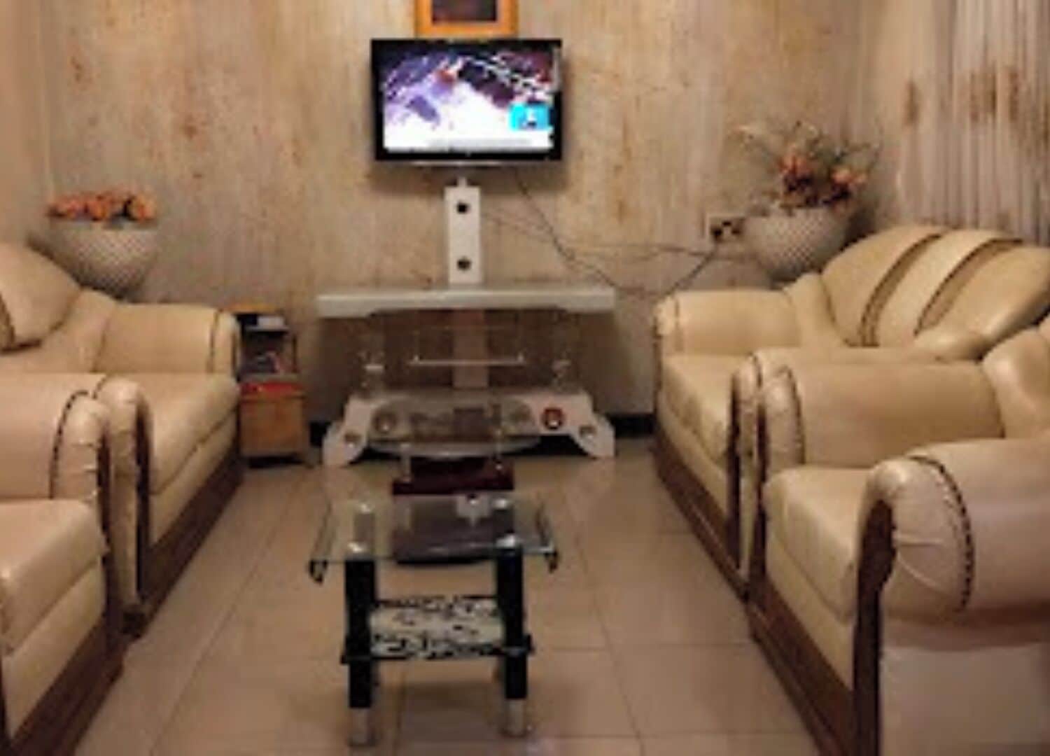 A living room with two beige leather sofas, a glass coffee table, a flat-screen TV mounted on the wall, and a small decorative plant.