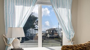 A room with a view of a house and a car through a window with light blue curtains. - Exquisitely Maintained Cottage Inside and Out, Open Layout, One Block to Beach,  *Pet Friendly* (Rockaway Beach)