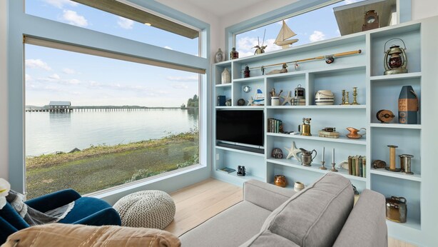 A modern living room with a large window overlooking a body of water, a flat-screen TV, and a collection of nautical-themed decor. - Bayfront Tiny Home - Tillamook Bay/Garibaldi (Garibaldi)