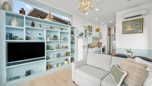 A living room with a TV, light blue shelves, a sofa, and a kitchen in the background. - Bayfront Tiny Home - Tillamook Bay/Garibaldi (Garibaldi)