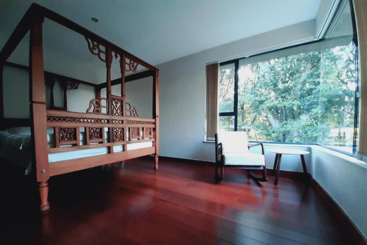 Una habitación con una cama de madera, una silla blanca y una mesita pequeña. Se ve una ventana grande con vistas a árboles.