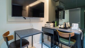 Une chambre d’hôtel moderne équipée d’un téléviseur à écran plat fixé au mur, d’un petit bureau avec une chaise et d’un coin cuisine compact.