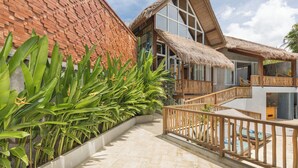 Un complexe hôtelier doté d’un bâtiment à toit de chaume, d’un balcon avec des balustrades en bois et d’un sentier bordé de plantes tropicales.