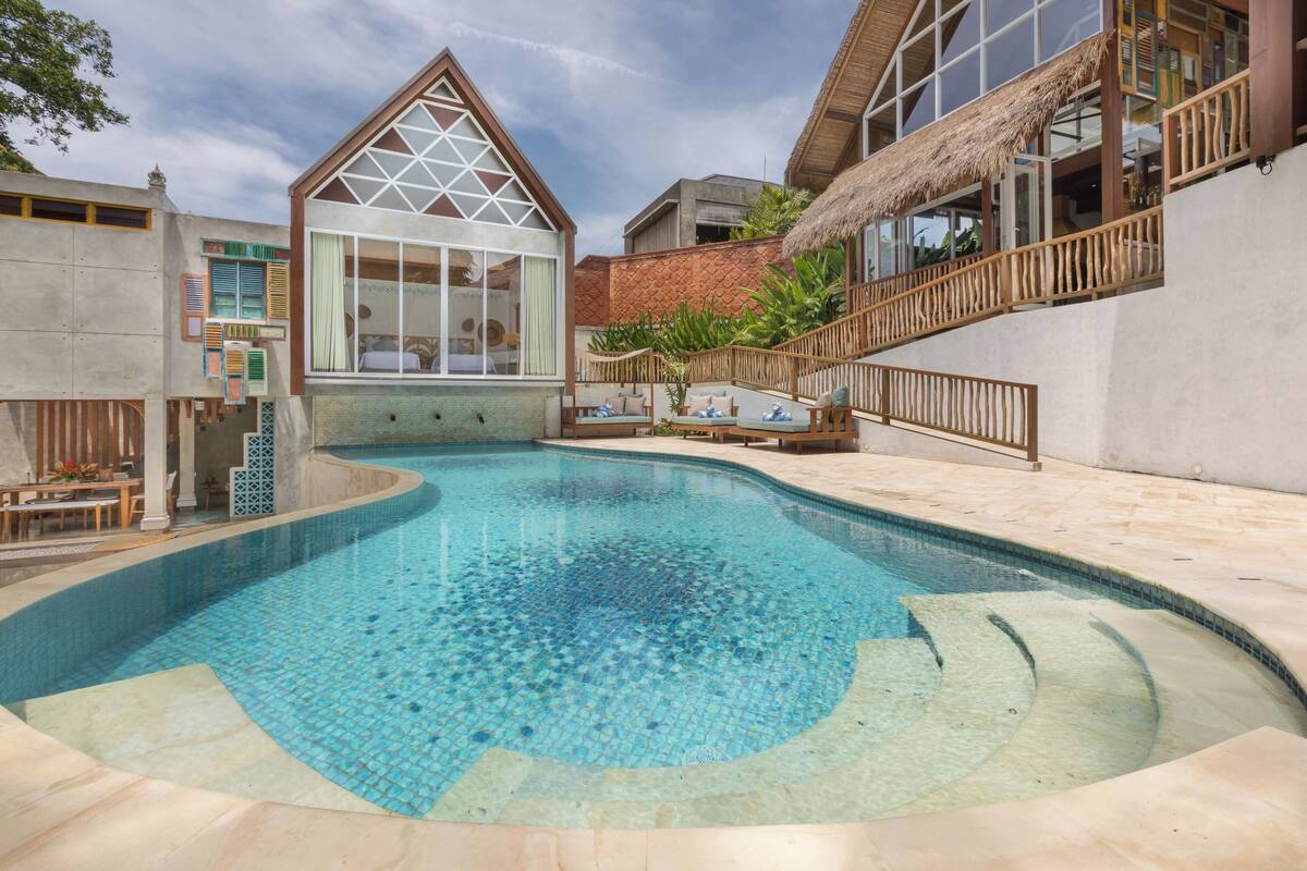 Una piscina con un diseño geométrico de azulejos, rodeada por un patio de concreto y una terraza de madera con techo de paja.