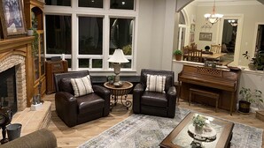 A living room with leather chairs, a fireplace, a piano, and a dining area visible through an archway.